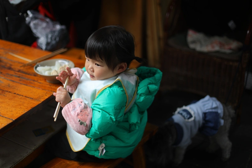 一个小女孩穿着一件绿色夹克坐在木制桌子上 用筷子从一碗面条里夹着吃东西。