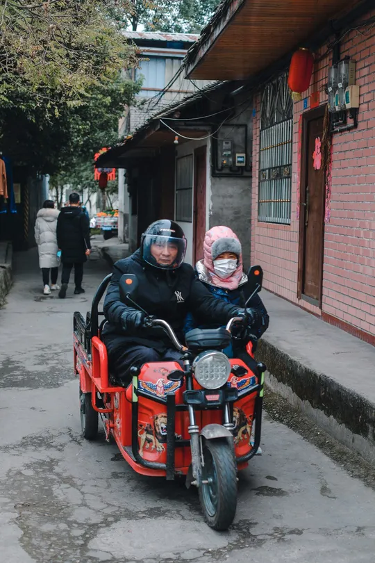 一个男子和女子带着一个小孩子骑着红色的摩托车沿着狭窄的街道行驶。