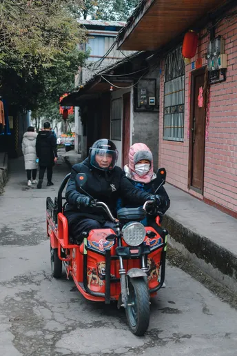 一个男子和女子带着一个小孩子骑着红色的摩托车沿着狭窄的街道行驶。
