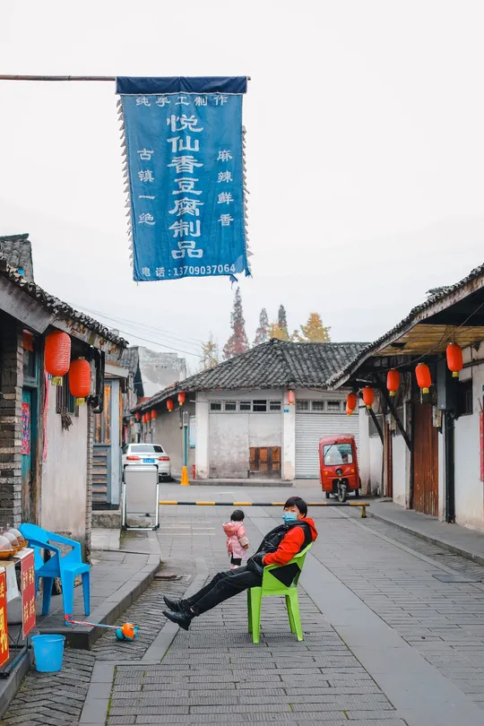 一个小孩子坐在街上的绿色椅子上 周围有建筑物和旗帜。