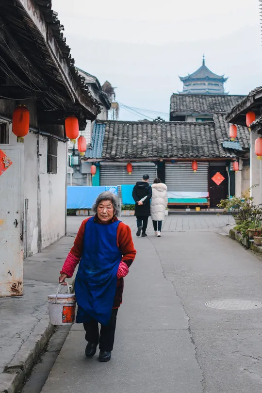 一个老人在村庄的巷子里拿着一个桶走下 周围有中国建筑和灯笼。
