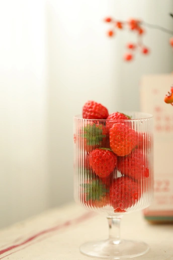 一个装满红果的透明玻璃碗 放在一张桌子旁边 旁边有一个小花瓶。