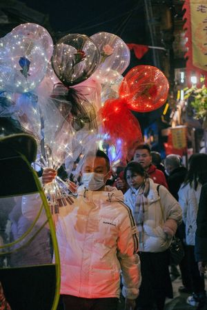 一个戴口罩的男子站在一个行人穿行、挂满彩球的街道中间。