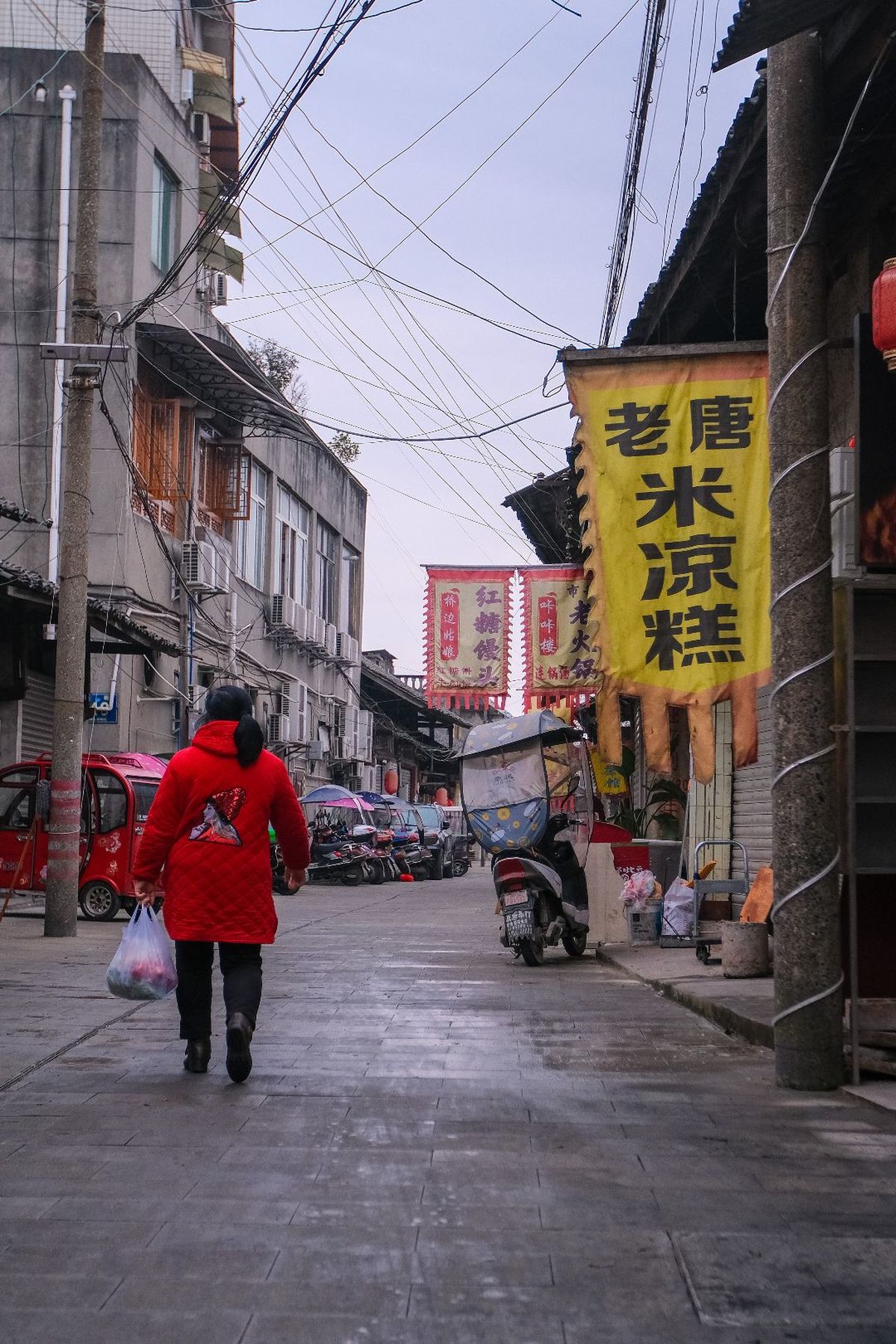 一个人穿着红夹克在亚洲城市的一条狭窄巷子里行走 周围有建筑物和一个带着包的男人。