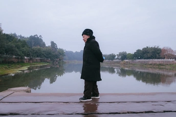 一个站在雨中 前方有类似湖的水体 手里拿着伞的男人。