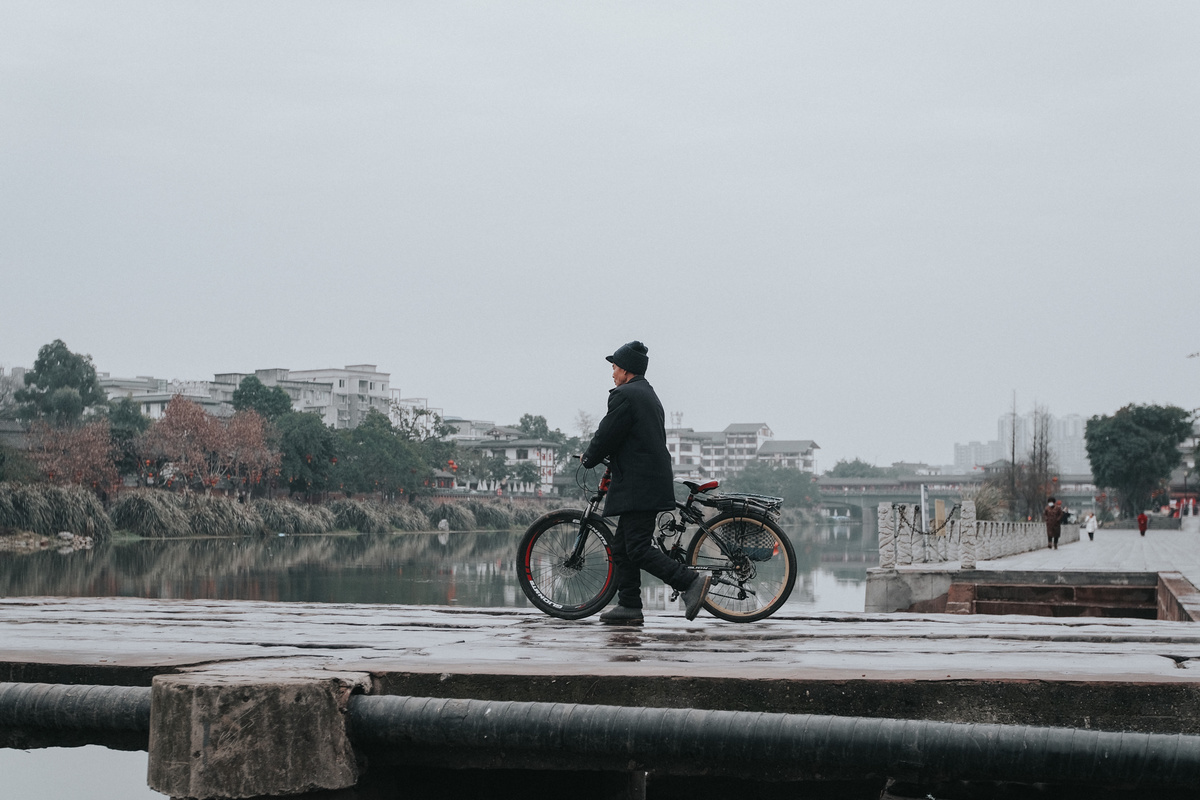 一个人在雨中撑着伞 步行推着自行车在码头附近的水边。