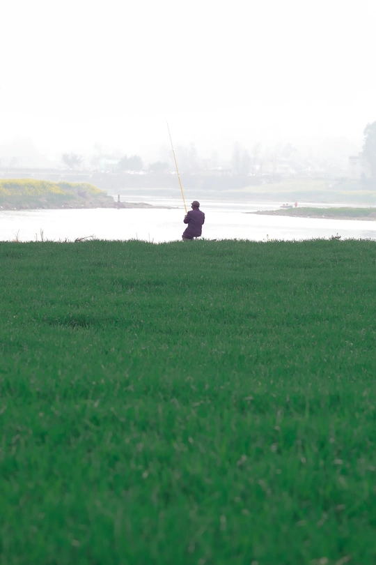 一个人坐在草地上 旁边有一个人在水中钓鱼 远处有一个人在风筝上钓鱼。
