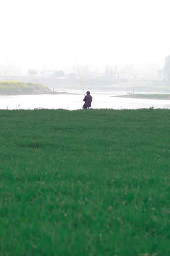 一个人坐在草地上 旁边有一个人在水中钓鱼 远处有一个人在风筝上钓鱼。