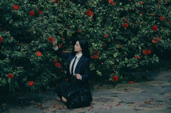 一位穿着黑色西装的女人跪在地上 手里拿着一个黑色袋子 站在开满红花的灌木丛前。
