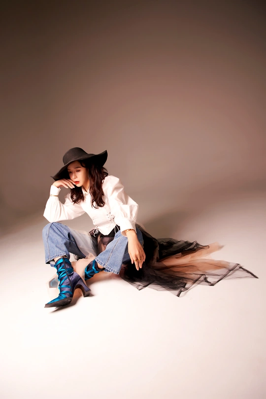 一位穿着白色衬衫、黑色牛仔帽、牛仔裤和靴子的年轻女子坐在地板上。