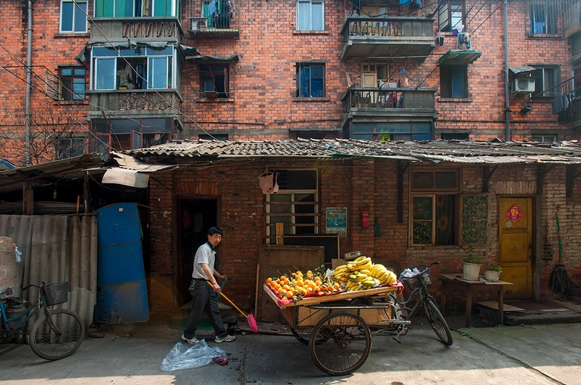 一个推着满载香蕉和其他水果蔬菜的小推车的人走在公寓楼前的一条街上。