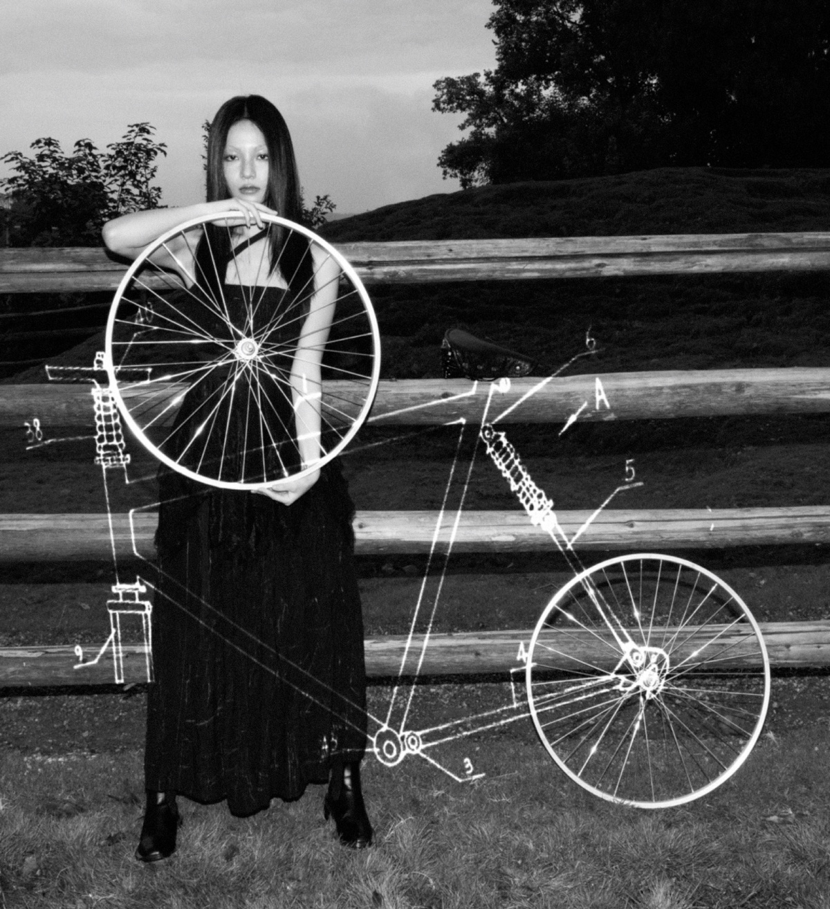 一张黑白照片 一位穿着连衣裙的女人站在自行车前轮前面 旁边有一道围墙。