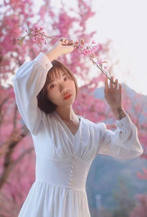 一位穿着白色连衣裙的女士在盛开的花树下拿着粉色花朵。