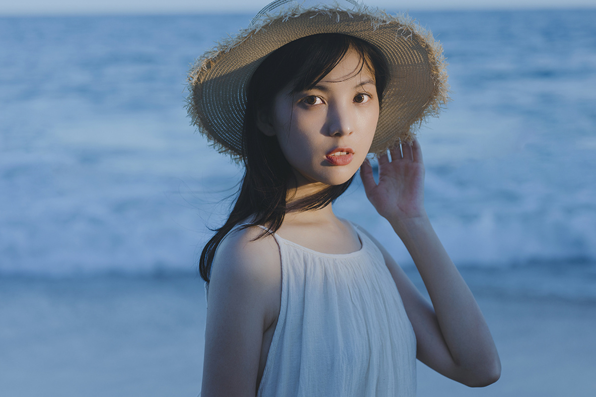 一位穿着白色连衣裙和草编帽子的年轻女子在海滩上靠近海洋摆姿势