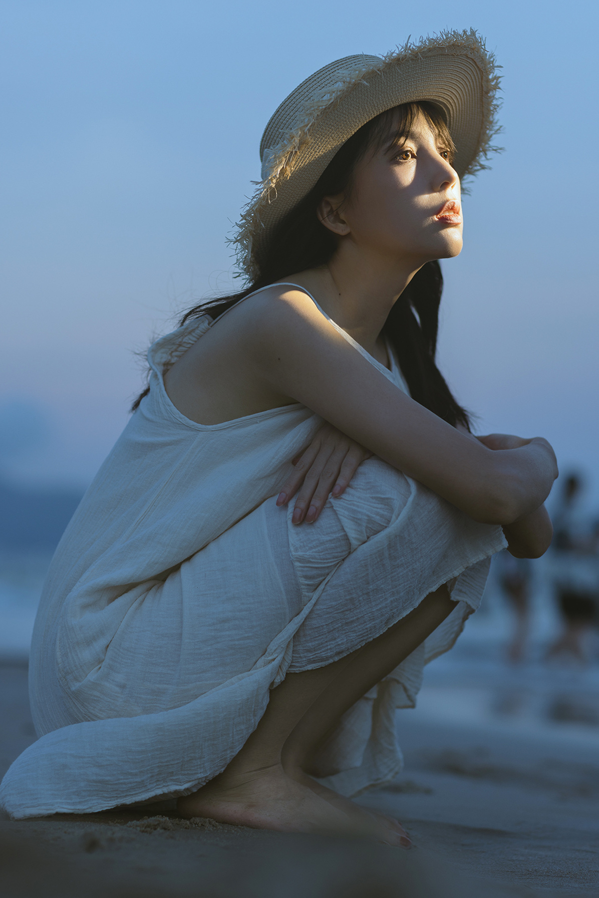 一位穿着白色连衣裙和草编帽子的年轻女子坐在海滩附近的水边