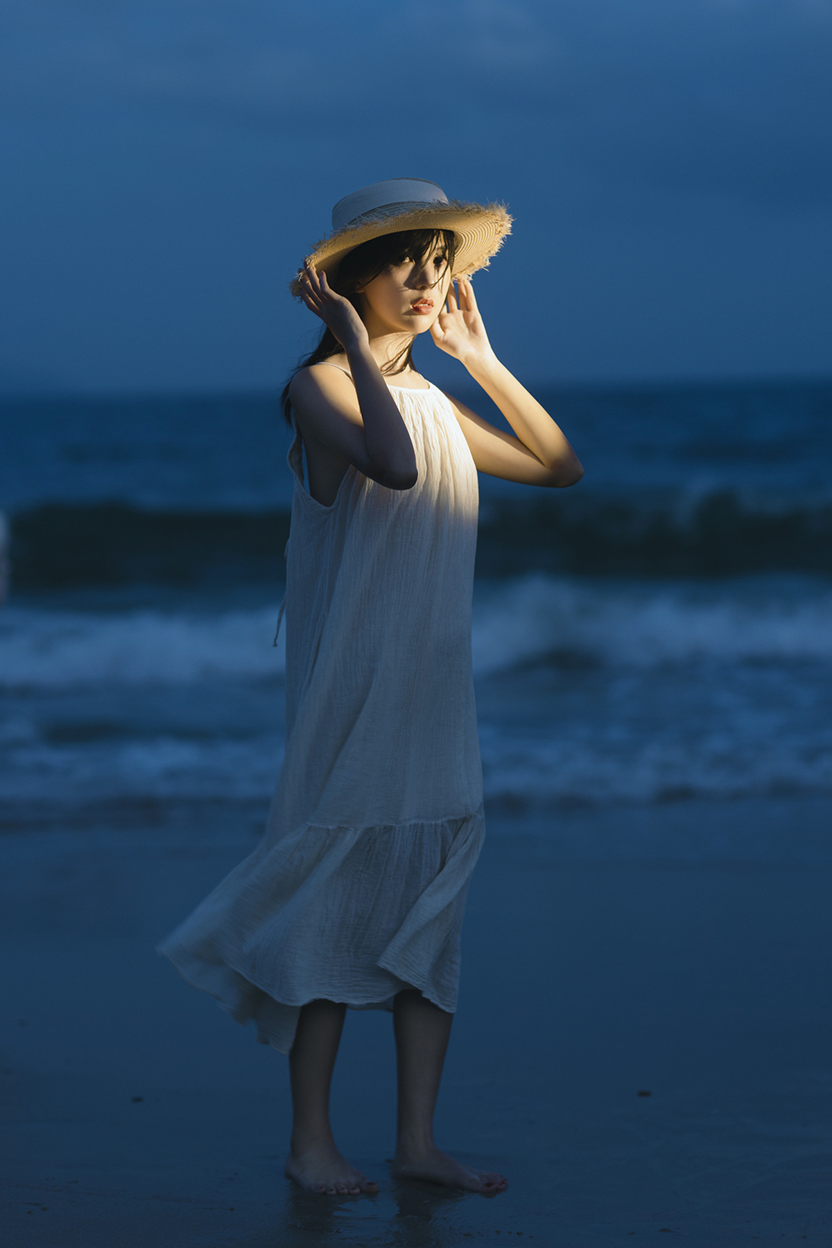 一个穿着白色连衣裙和草编帽子的年轻女子站在夜晚的海滩上 靠近海洋。
