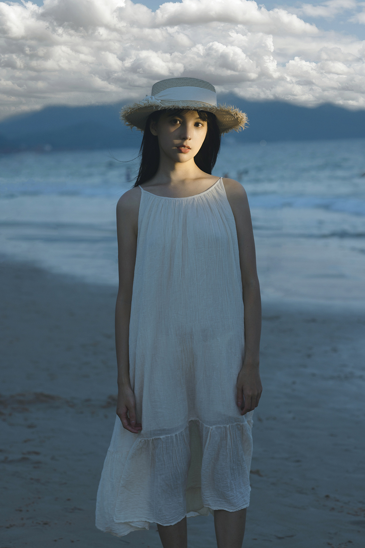一位穿着白色连衣裙和草编帽子的年轻女子站在海滩附近的海边