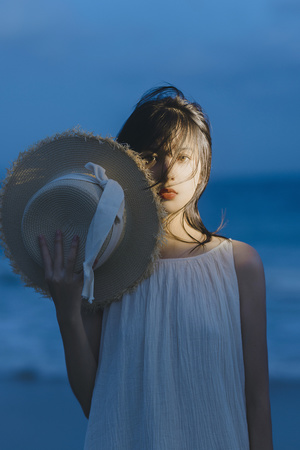一位穿着白色连衣裙和一顶大草帽的年轻女子在海滩上靠近海洋摆姿势