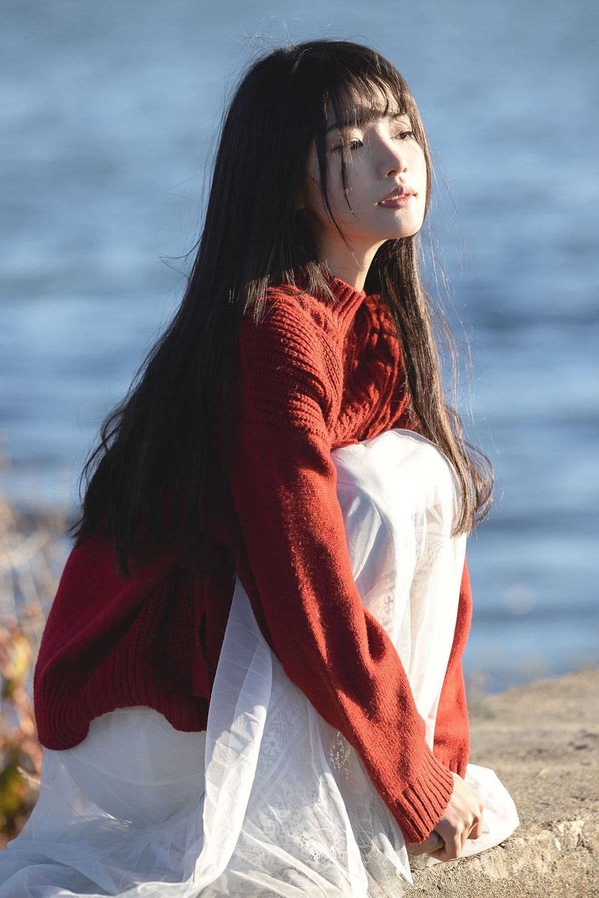 一个穿着红色毛衣和白色裙子的年轻女子坐在海边的一块岩石上