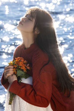 一位穿着红色毛衣的年轻女子站在水边 手里拿着花。