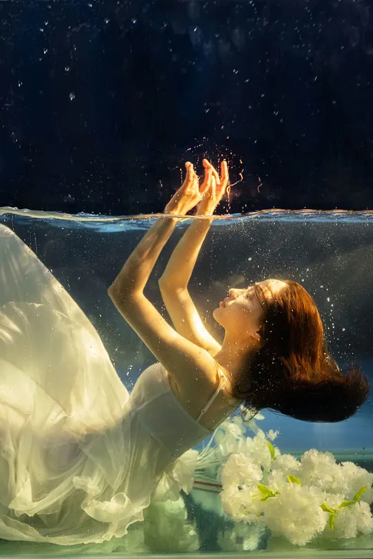 在水族馆中 一位穿着白色裙子的女人在水下 周围有花。
