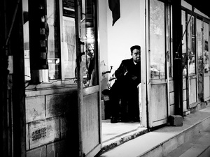 一张黑白照片 一位男子坐在人行道上的一把椅子上 这家商店就在他面前。