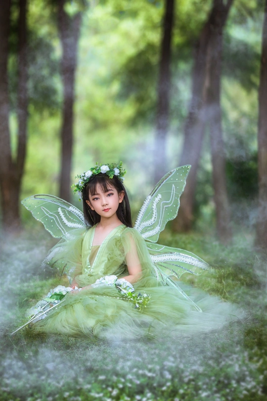 一个打扮成长着绿色翅膀的美丽仙女的小女孩坐在树林里
