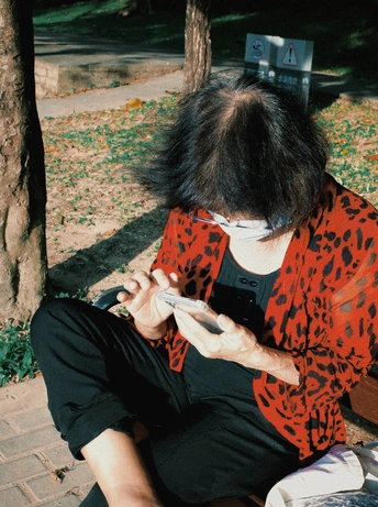 一位妇女坐在人行道旁的一棵树下长椅上 正在看手机并持有一把剪刀。