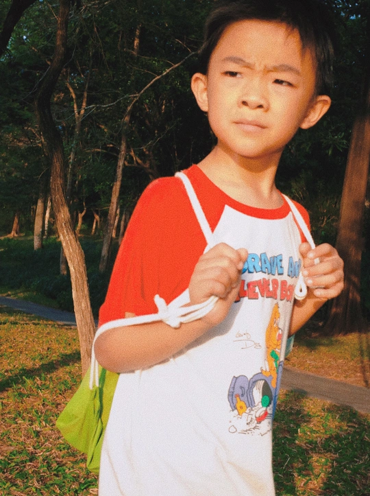 一个穿着红白相间T恤的小男孩站在公园里 手里拿着一块小滑板。