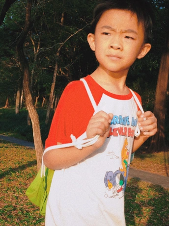 一个穿着红白相间T恤的小男孩站在公园里 手里拿着一块小滑板。