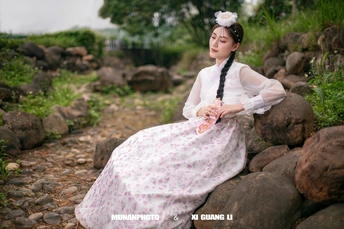 一位穿着传统服装的年轻女子坐在一条小溪旁的岩石上 手里拿着一朵白色花。