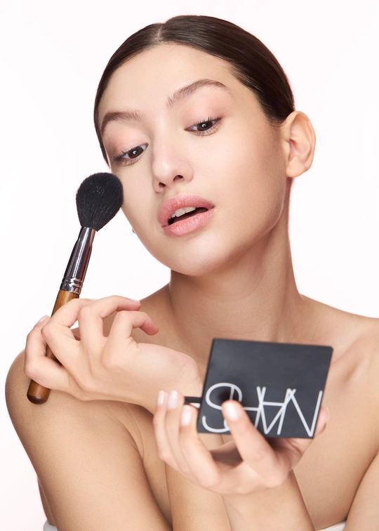 一位美丽的年轻女子正在用刷子刷她的脸颊并往脸上扑粉。