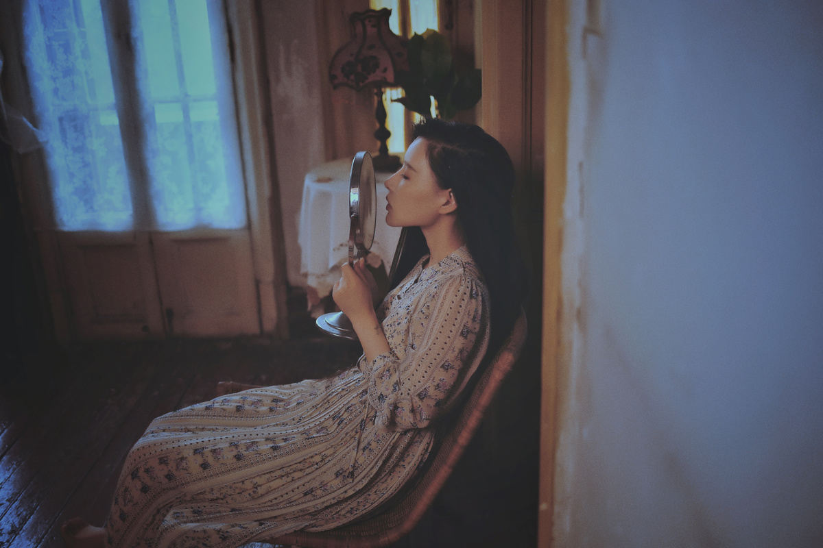 一个穿着连衣裙的年轻女子坐在一个有窗户和镜子的房间的椅子上