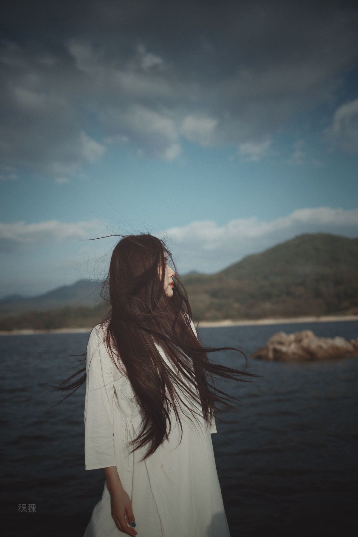 一位长发女子穿着白色长裙 站在水边吹风。