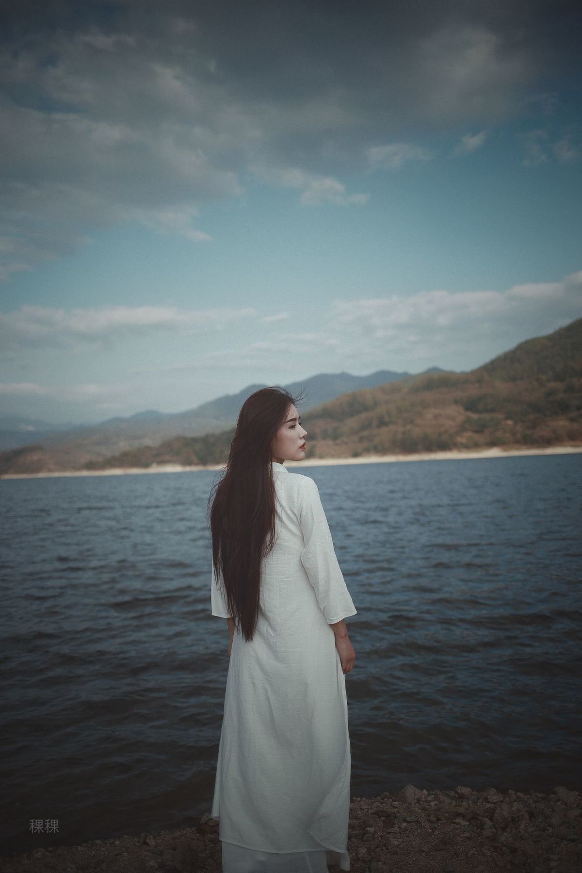 一位穿着白色连衣裙的女子站在水边 眺望着大海。