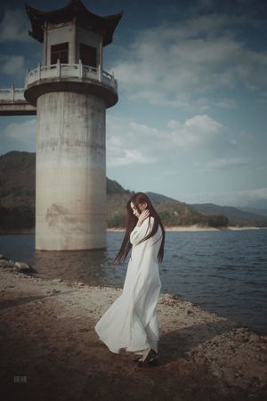 一位穿着白色连衣裙的美女站在水边 背景中有灯塔。