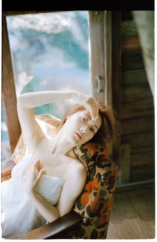 一个穿着白色连衣裙的女人坐在木椅上 面前有一扇窗户。