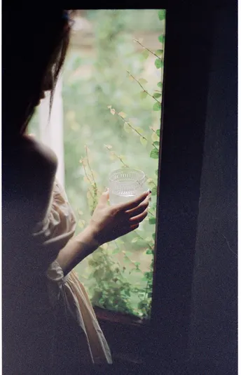 一位女士拿着一个玻璃窗 凝视着窗外的花。