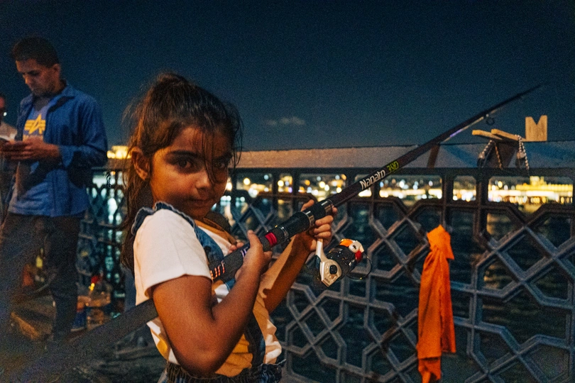 一个年轻女孩站在夜晚的码头上 手里拿着钓鱼竿 而一个男子在水中钓鱼。