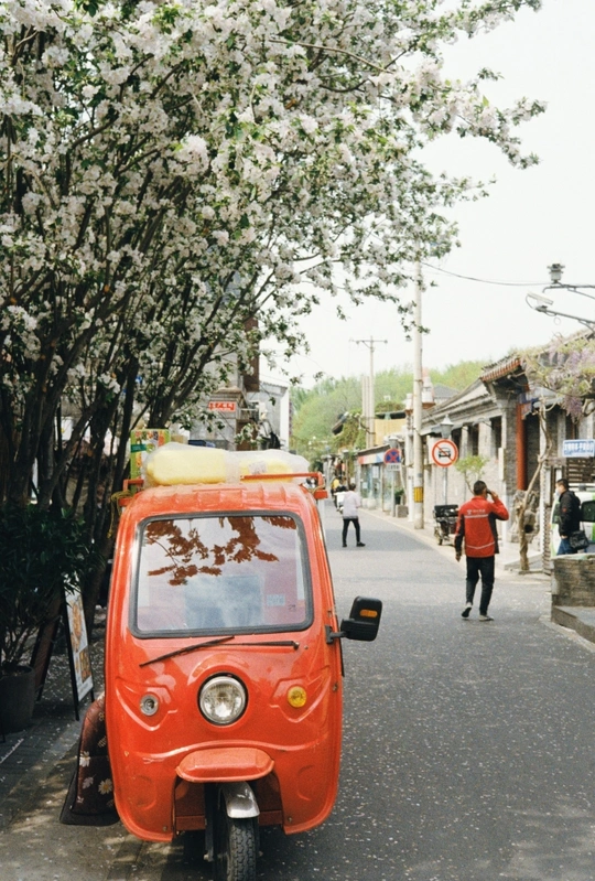 一辆橙色汽车沿着街道行驶 路边停着一辆小摩托车 路上有行人行走。