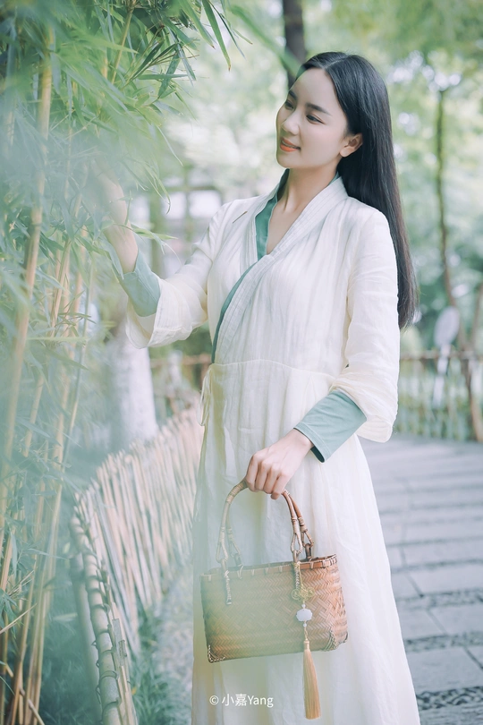 一个穿着传统白色连衣裙、拿着包的漂亮年轻女子在竹林中行走