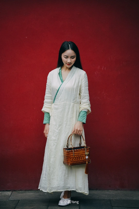 一位身穿白色长裙、提着篮子的年轻女子靠着红墙站着
