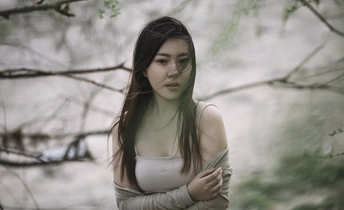 一位穿着白色连衣裙的年轻女子站在森林中 周围有树木和水。