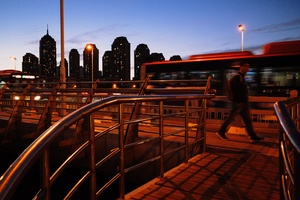 一个人在黄昏时走过一座桥 背景是城市天际线 夜晚有一辆公交车经过。