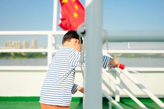 一个小男孩举着中国国旗站在一艘船的甲板上 眺望着水面。