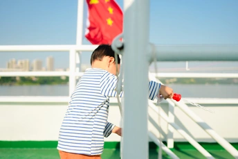 一个小男孩举着中国国旗站在一艘船的甲板上 眺望着水面。