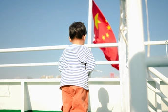 一个小男孩站在一艘船的甲板上 看着从栏杆上飘过的红旗。