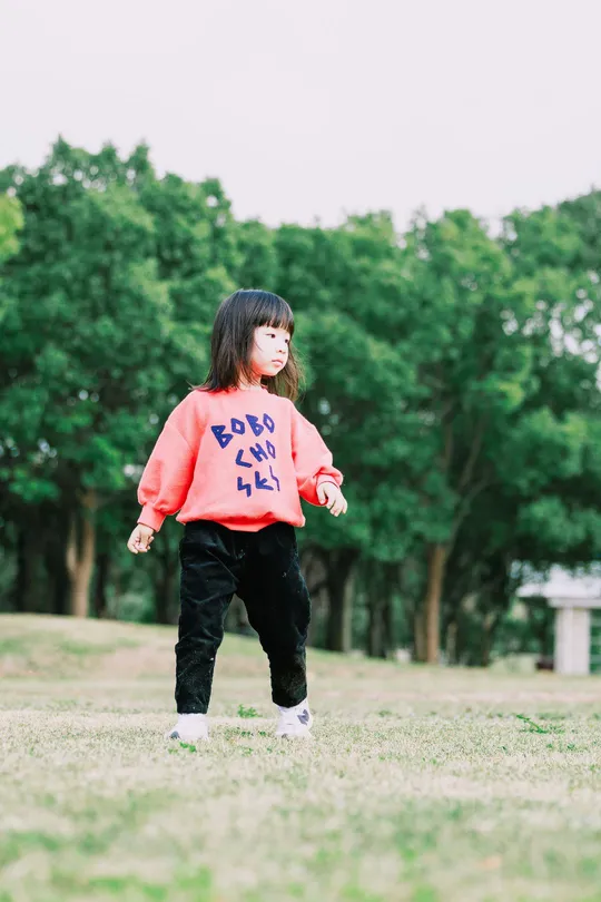 一个小女孩穿着一件写着“小联盟”的衬衫站在公园的草地上 一个小孩子站在她后面。