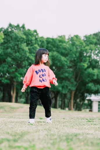 一个小女孩穿着一件写着“小联盟”的衬衫站在公园的草地上 一个小孩子站在她后面。
