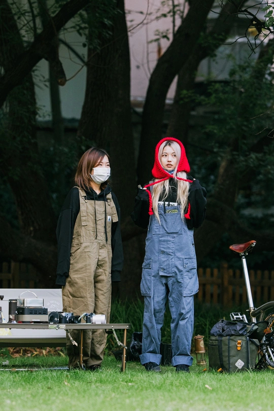 一个戴红面具的男子站在一个穿工作服的年轻女孩旁边 背景是一个有摩托车的院子。